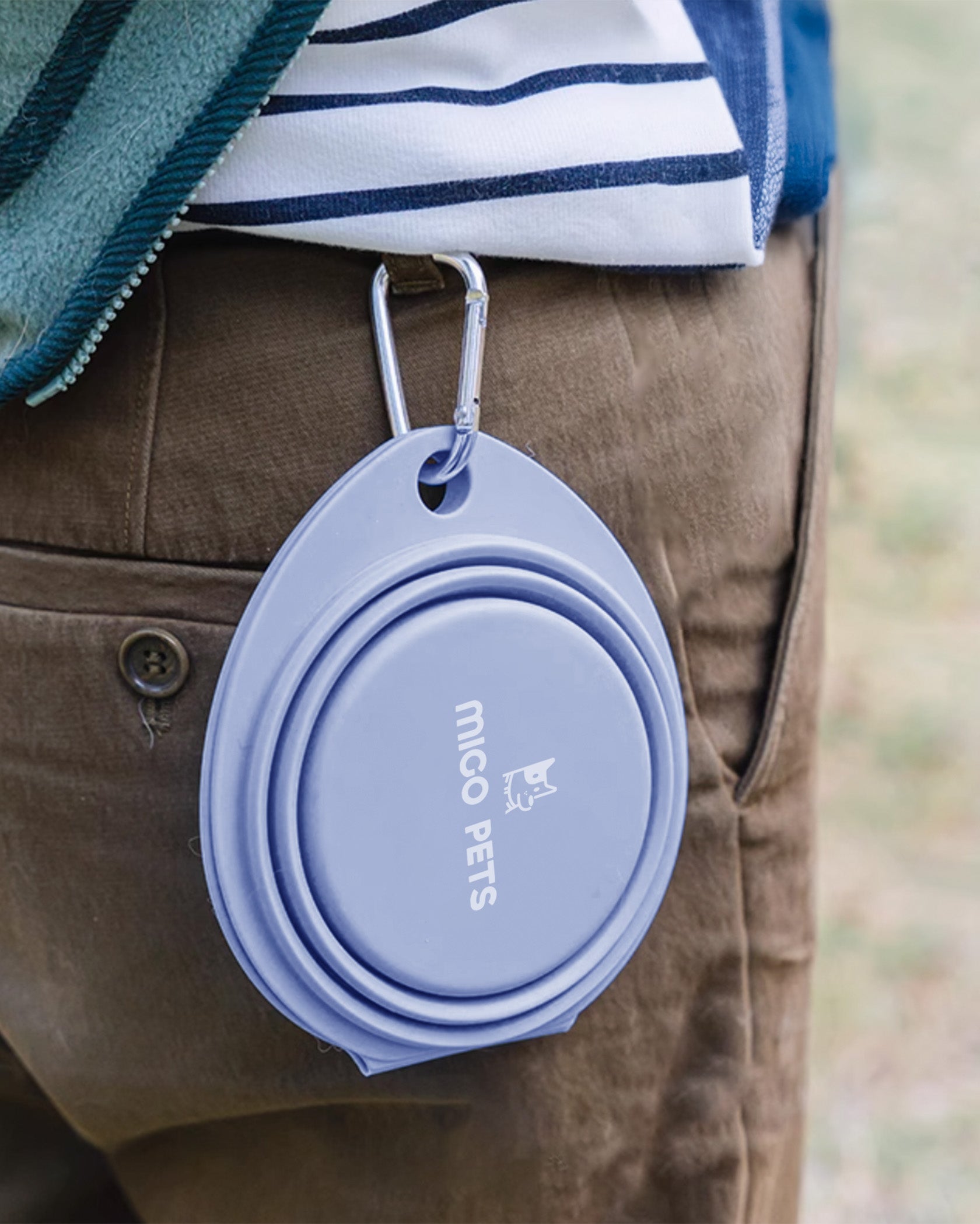 A folded double dog bowl in soft dusty blue, clipped onto a belt loop with a carabiner, ideal for travel and outdoor use.
