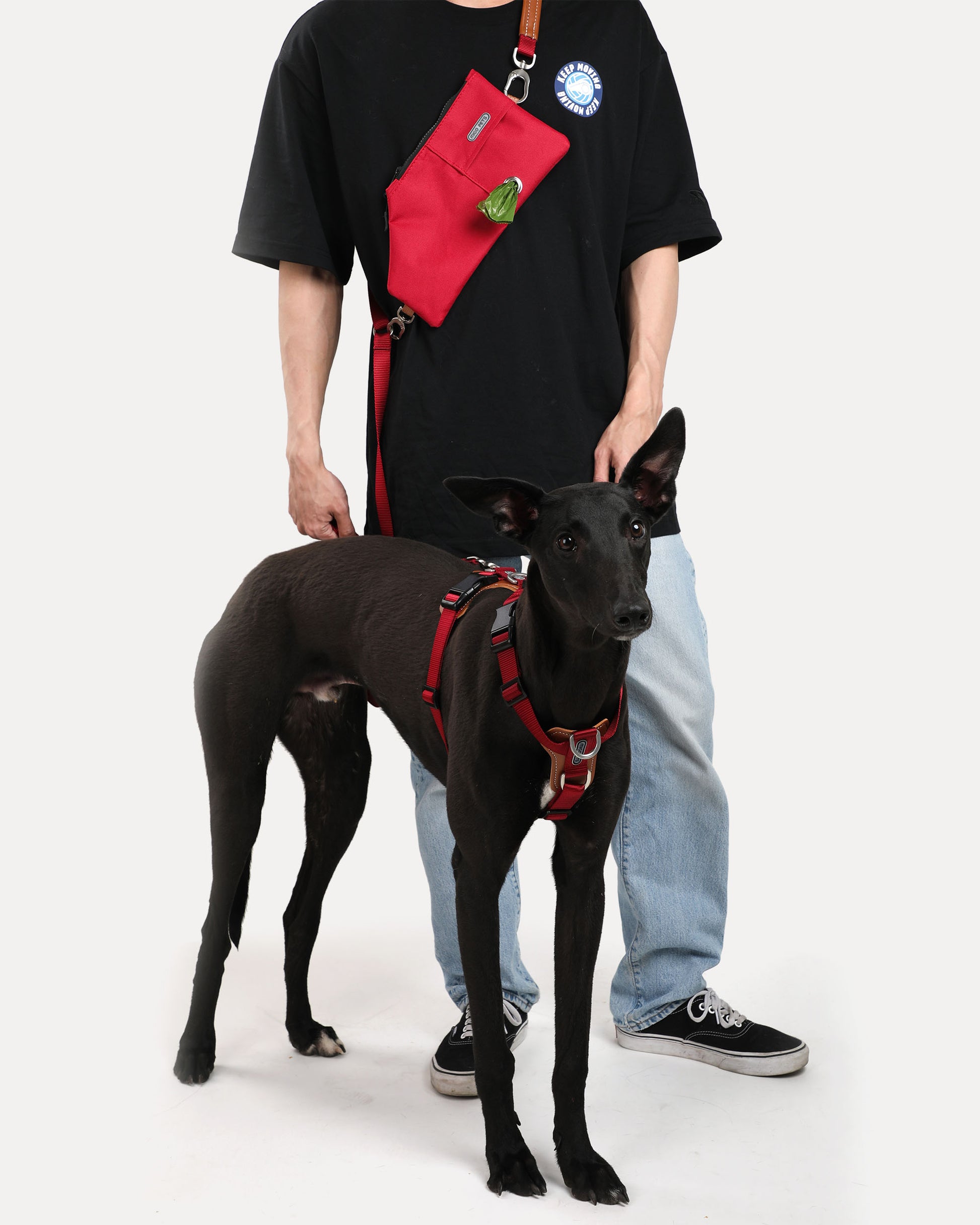A dog in a crimson red step-in adjustable dog harness, with the owner using a hands-free crossbody dog leash and a crossbody travel pouch.